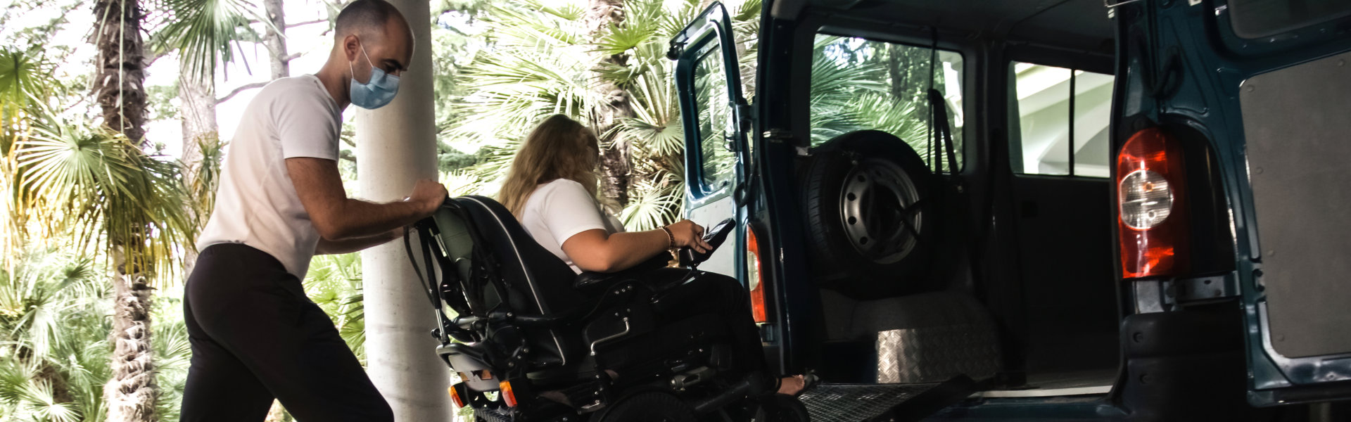 Man on wheelchair using accessible vehicle with ramp for transportation with driver helping.